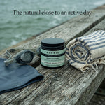 A jar of Kloris Rapid Comfort Body Butter sits on a weathered wooden dock beside a striped towel, swim goggles, and a dark rubber pad, with water in the background and the text 'The natural close to an active day.'.