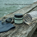 A jar of Kloris Rapid Comfort Body Butter sits on a weathered wooden dock beside a striped towel, swim goggles, and a dark rubber pad, with water in the background and the text 'The natural close to an active day.'.