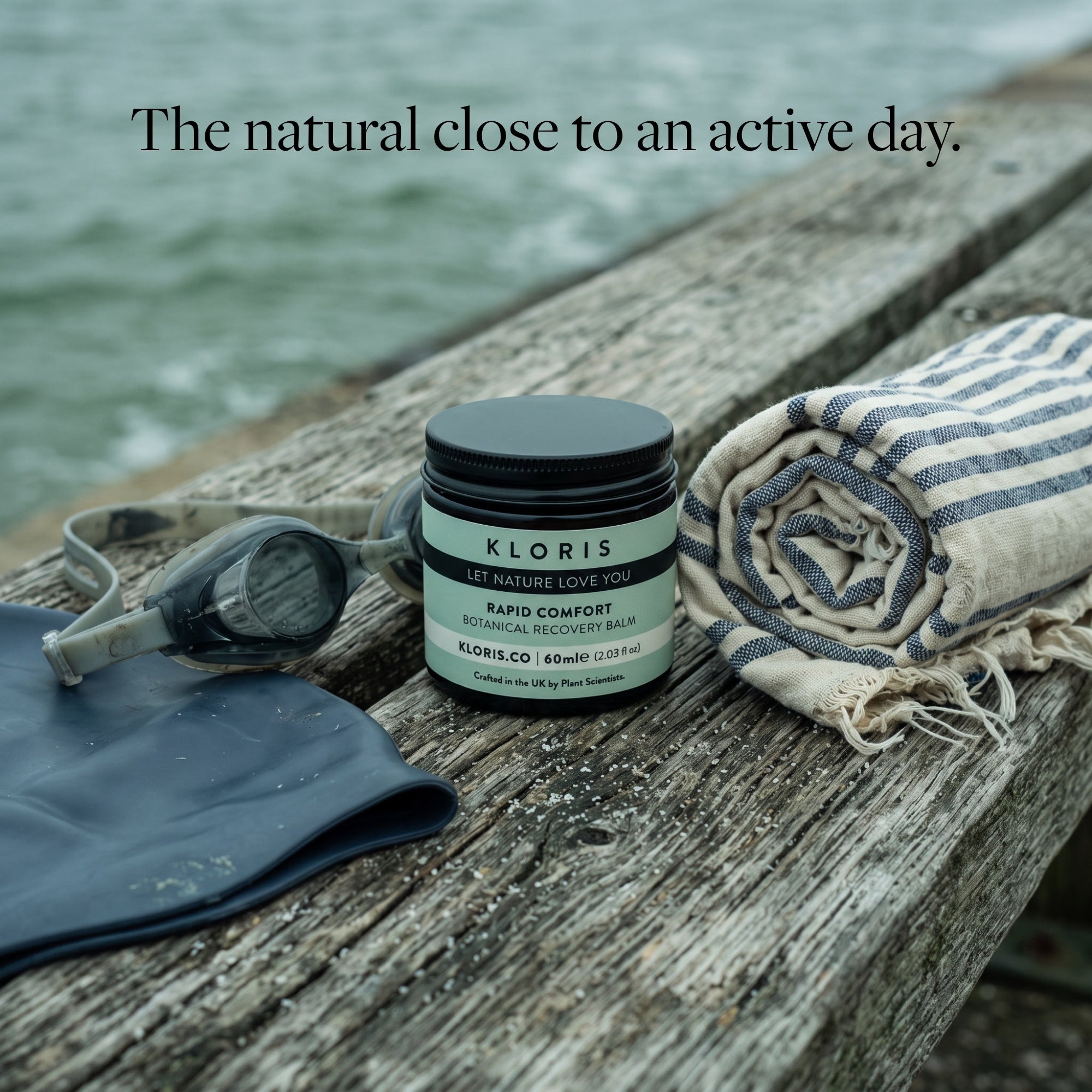 A jar of Kloris Rapid Comfort Body Butter sits on a weathered wooden dock beside a striped towel, swim goggles, and a dark rubber pad, with water in the background and the text 'The natural close to an active day.'.