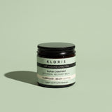 A jar of Kloris Rapid Comfort Botanical Recovery Balm on a solid sage green background, featuring a black lid and a black and white label.