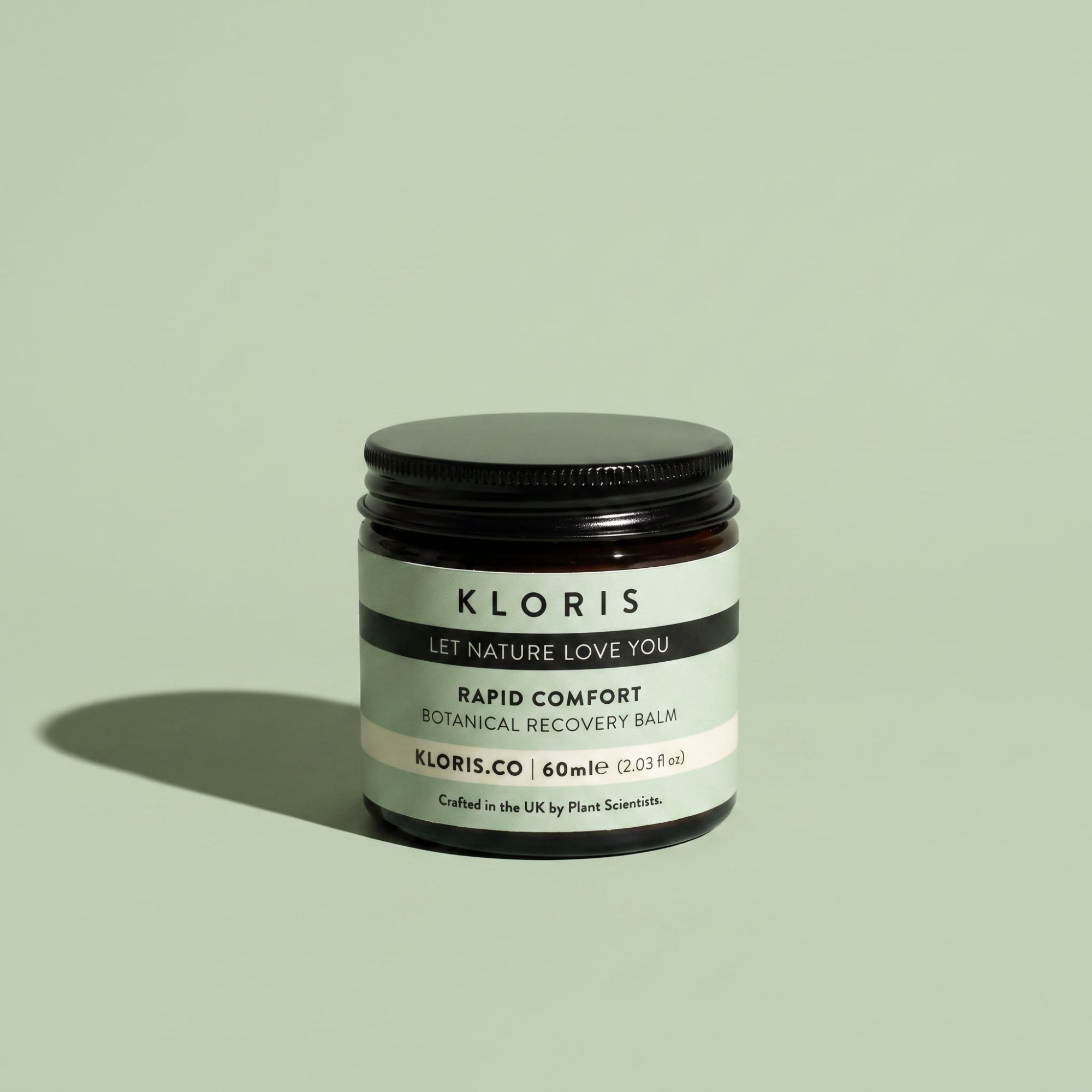 A jar of Kloris Rapid Comfort Botanical Recovery Balm on a solid sage green background, featuring a black lid and a black and white label.