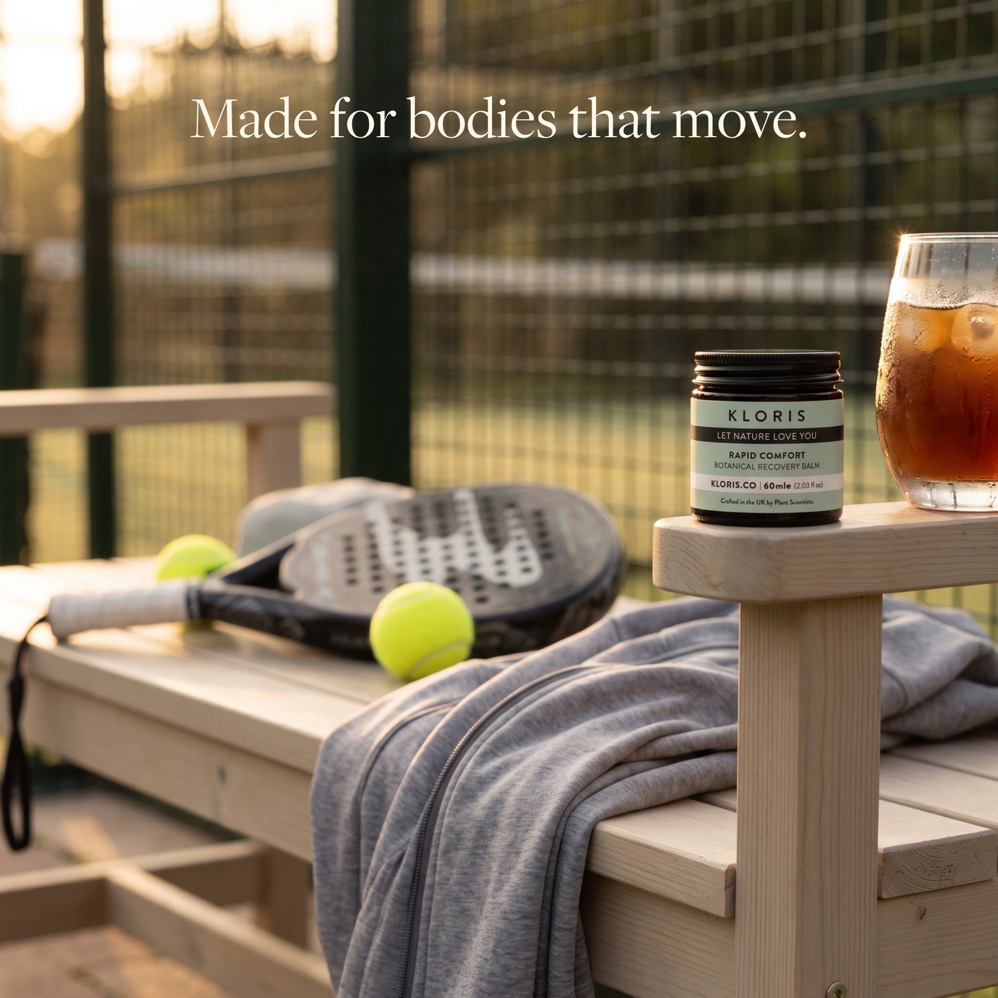A lifestyle shot of a Kloris balm jar on a wooden bench next to a glass of iced coffee, with a tennis racket and tennis balls in the background, under the text 'Made for bodies that move.'.