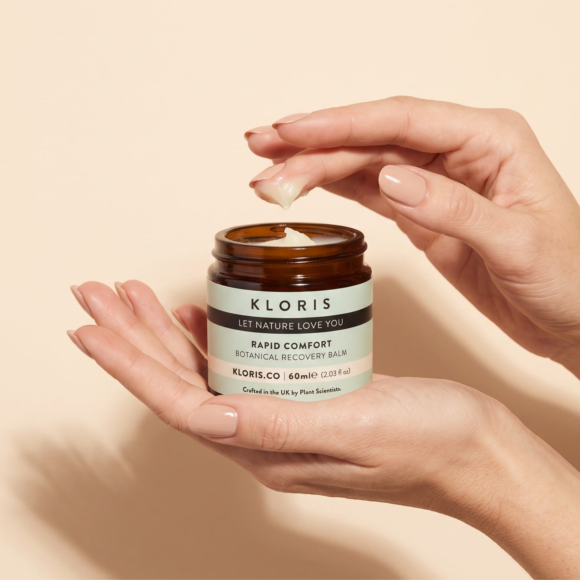 A person's hands holding an amber glass jar of Kloris Rapid Comfort Botanical Recovery Balm, with a dollop of white balm being lifted from the jar against a warm beige background.