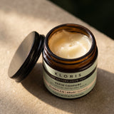 An open amber glass jar of Kloris Rapid Comfort Botanical Recovery Balm with its black lid resting beside it, set on a textured beige stone surface in warm natural light.