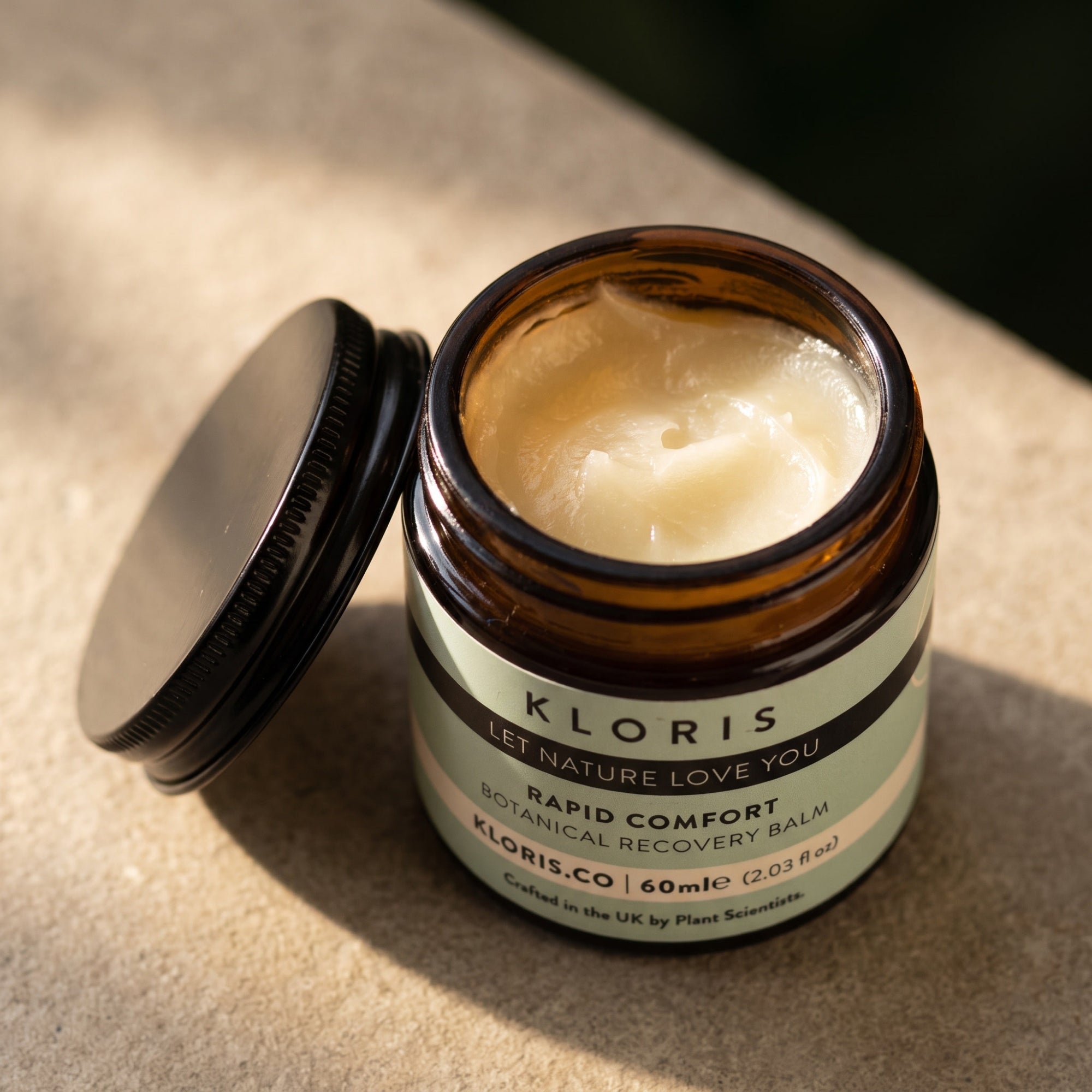 An open amber glass jar of Kloris Rapid Comfort Botanical Recovery Balm with its black lid resting beside it, set on a textured beige stone surface in warm natural light.