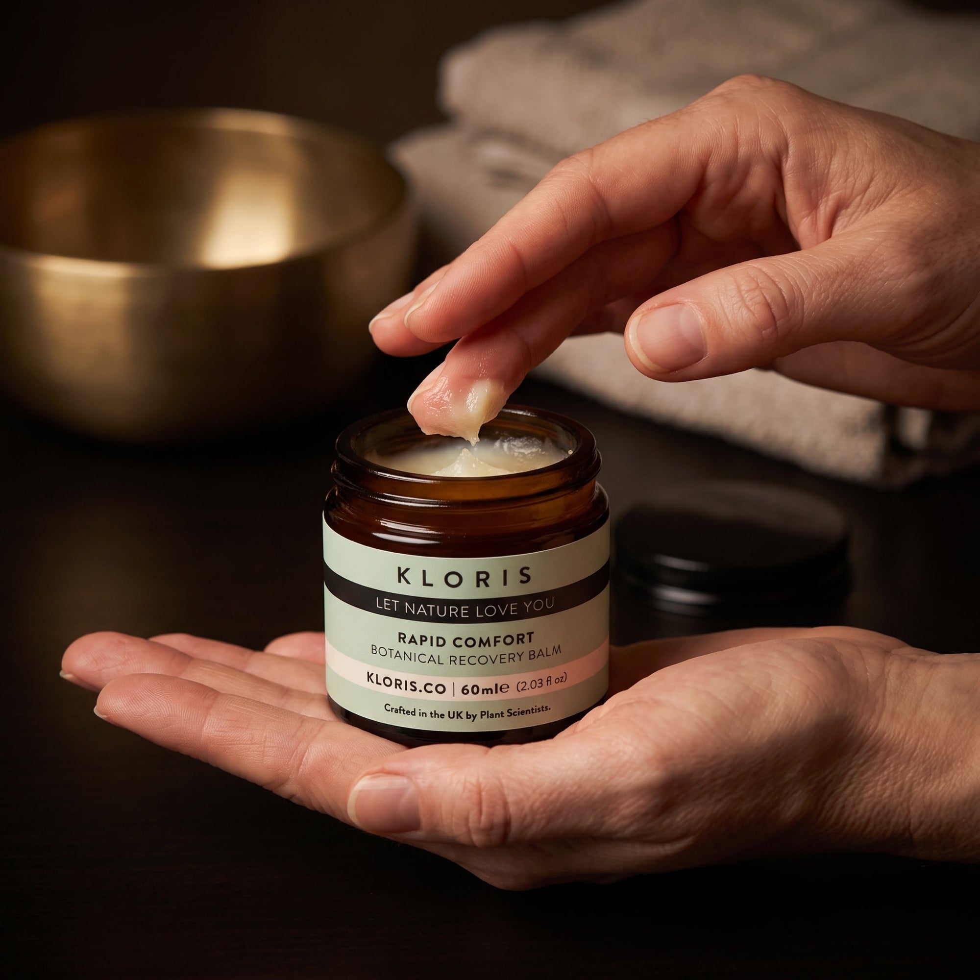A close-up of a person's hands holding an amber glass jar of Kloris Rapid Comfort Botanical Recovery Balm, with one hand lifting the lid to show the balm, set against a dark background with a gold bowl and a towel.
